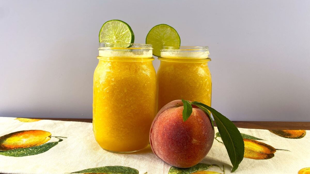 Two peach daiquiries in mason jars, with limes on their rims and a ripe peach for company