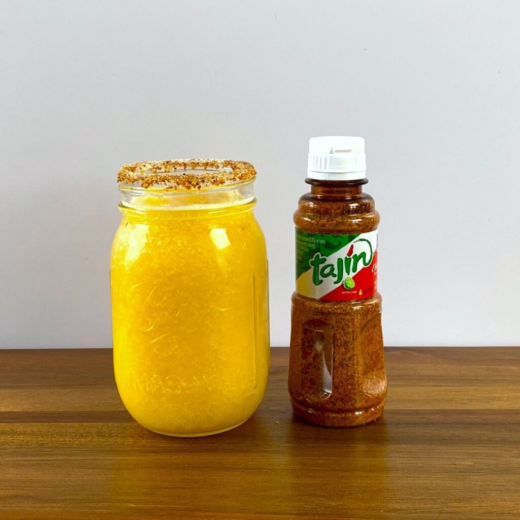 A chili lime spice rimmed peach daiquiri beside a bottle of Tajin spice blend