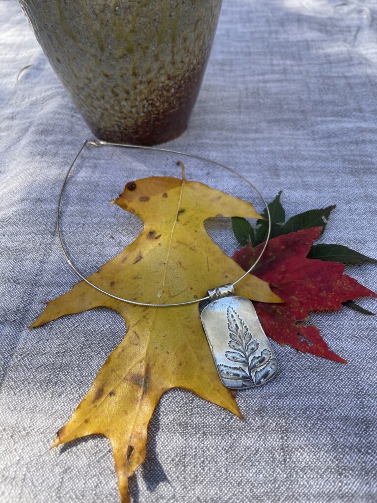 An artisanal metal bracelet with a plant design, resting on fall leaves.