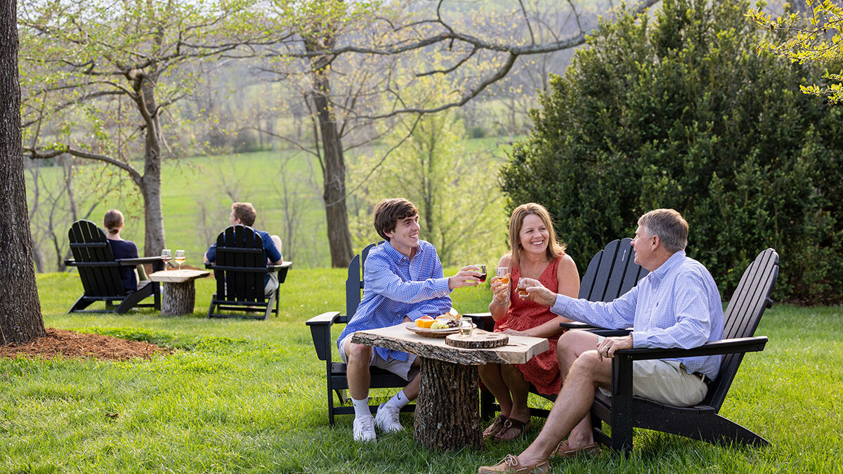 A family drinks wine and enjoys a charcuterie board outdoors, seated in Adirondak chairs.