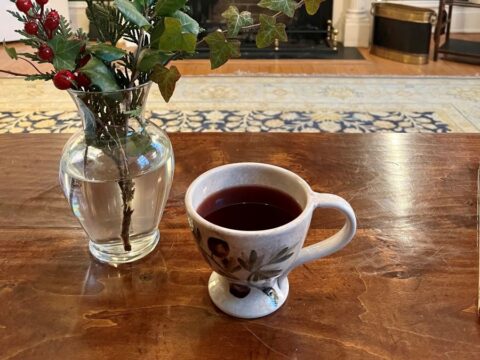 A cup of mulled wine on a table near some festive plants in a vase.