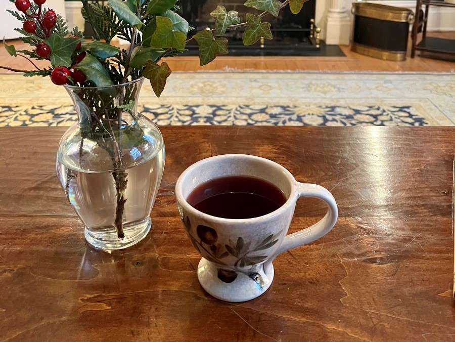 A cup of mulled wine on a table near some festive plants in a vase.