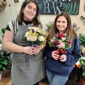 Two women holding flower arrangements, with a sign reading "Flower Bar" behind them