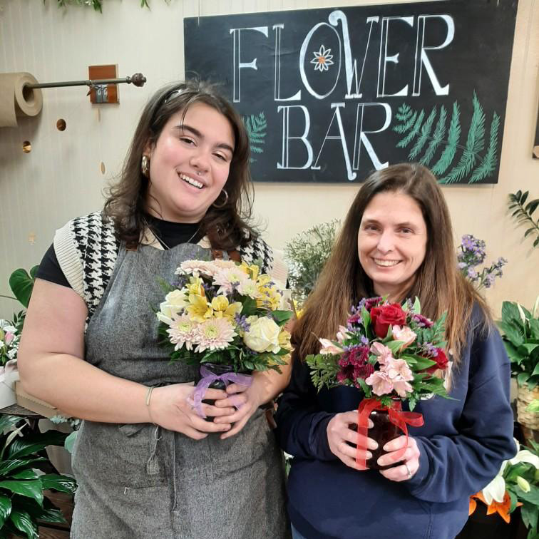 Two women holding flower arrangements, with a sign reading "Flower Bar" behind them