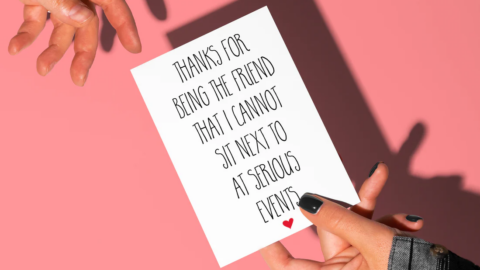 Two hands exchanging a card reading "Thanks for being the friend that I cannot sit next to at serious events"