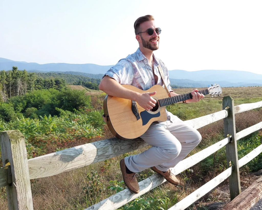 A guitarist in sunglasses sitting on a wooden fence outside